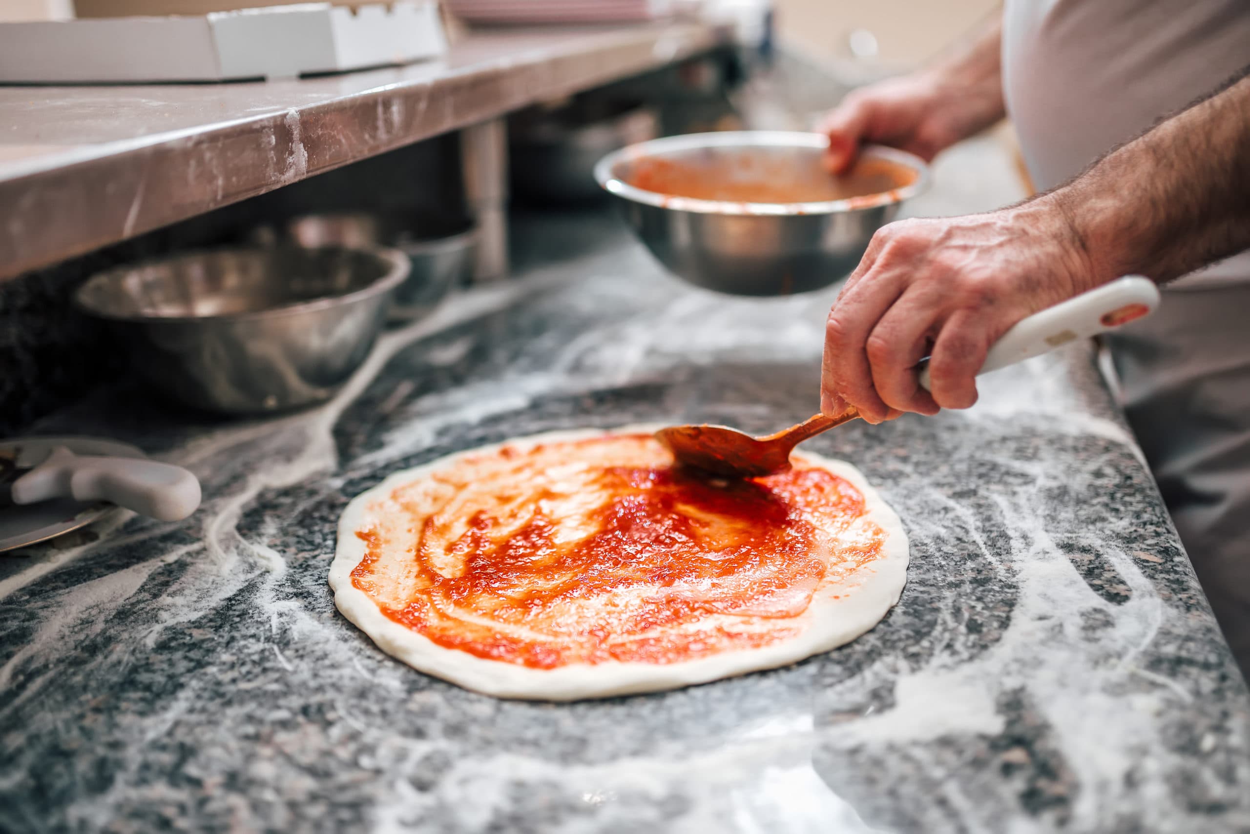 Tomatensauce wird mit einer Kelle auf Pizzateig verstrichen